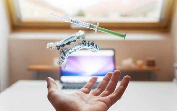 A photograph of a hand held out with transparent aligner trays and a whitening gel syringe seemingly floating above it in an office setting.