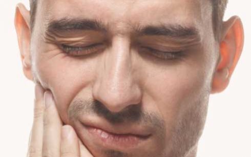 A close-up photograph of a man with his eyes closed, holding his hand to his jaw in clear expression of pain.