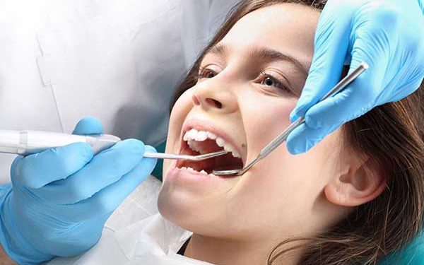 A close-up photograph of a young girl undergoing a dental examination while a dentist wearing blue gloves uses a dental mirror and explorer.