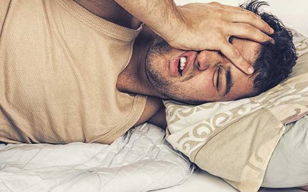 A photograph of a man lying in bed holding his head in pain, illustrating dental discomfort or a toothache.