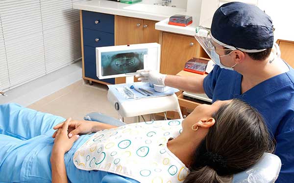 A photograph of a dentist wearing personal protective equipment pointing at a dental x-ray screen while explaining the treatment plan to a patient in a dental chair.