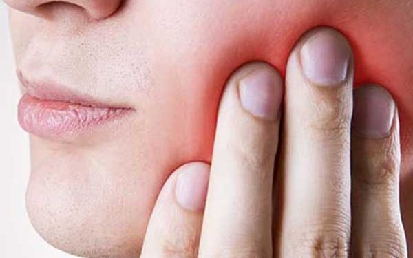 A close-up photograph of a person holding their hand to their cheek, experiencing facial and dental pain.