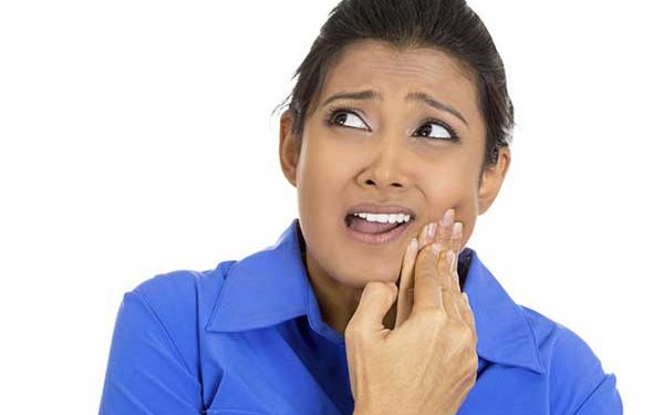 A photograph of a woman wearing a blue top, holding her hand to her cheek with an expression of discomfort or toothache.