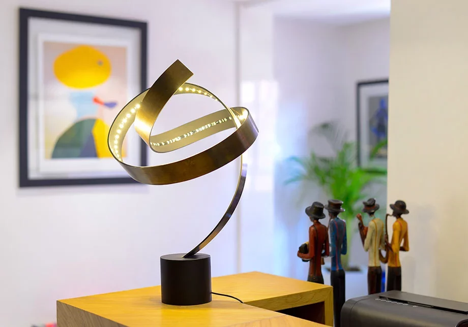A photograph of an artistic, illuminated metal ribbon sculpture acting as a desk lamp, resting on a wooden surface in a contemporary office interior.