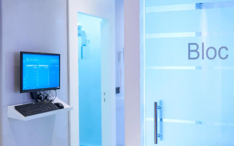 A photograph of a modern clinic hallway featuring a digital workstation monitor on the wall and a frosted glass door marked with the word "Bloc".