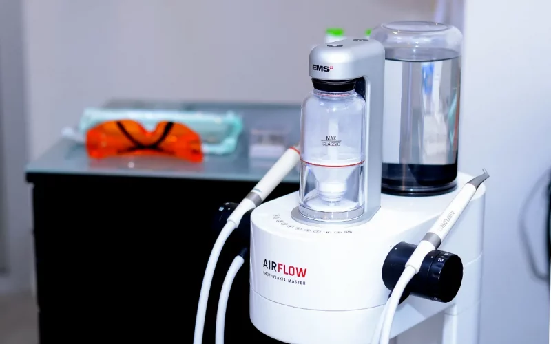 A close-up photograph of an EMS Airflow Prophylaxis Master dental cleaning unit with its powder chamber and hand pieces on a cart in a clinic.