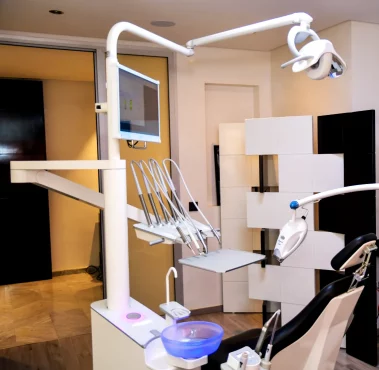 A photograph of a modern dental treatment room featuring an ergonomic dental chair, delivery unit, monitor, and overhead light fixture.