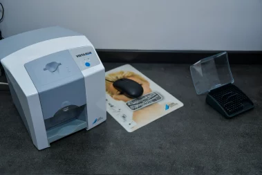 A close-up photograph of a Dürr Dental VistaScan scanner, a computer mouse on a branded pad, and an open storage box on a dark clinic counter.