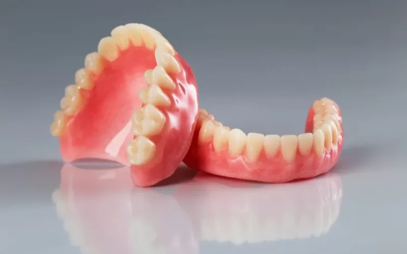 A photograph of complete upper and lower acrylic dentures resting on a reflective surface against a gradient background.