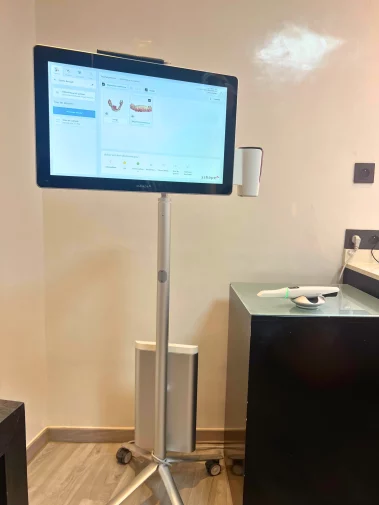 Mobile 3Shape digital scanning unit on a wheeled stand, displaying a patient's dental impression scans, with the intraoral scanner resting on a nearby cabinet.