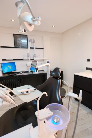 Close-up view of dental equipment including an LED sink basin, tool tray, overhead light, and a black dental chair, with a computer workstation in the background.