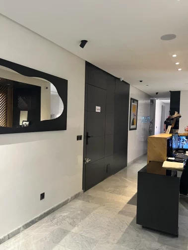 A modern interior hallway featuring a black designer mirror, a dark paneled exit door marked "Sortie," a reception desk, and marble tile flooring.