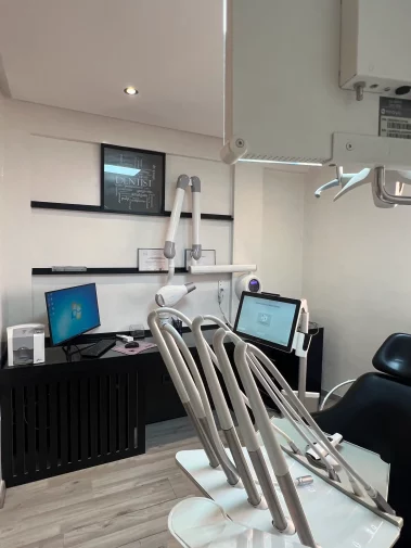 A modern dental treatment room featuring a black and white dental chair, digital computer screens, organized medical tool arms, and a framed "Dentist" word-art print on the wall.
