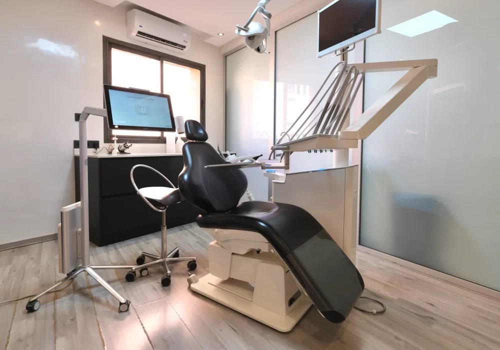 Modern dental treatment room with a black reclining dental chair, overhead light, monitor screens, and a dark cabinet workstation near a window.