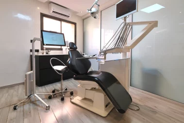 Modern dental treatment room with a black reclining dental chair, overhead light, monitor screens, and a dark cabinet workstation near a window.