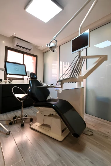 Modern dental treatment room with a black reclining dental chair, overhead light, monitor-mounted equipment arm, and a wall-mounted display screen.