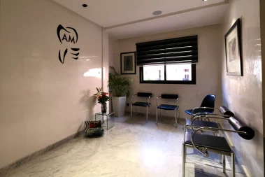 An interior photograph of the AM Dental Care waiting room featuring the practice logo on a white wall, minimalist black and chrome chairs, marble flooring, and a window with dark blinds.