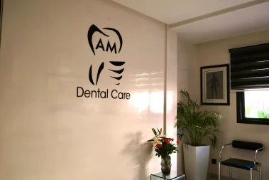 An interior photograph of a dental clinic waiting area featuring the "AM Dental Care" logo on a white wall, a potted plant, fresh flowers, and minimalist seating.