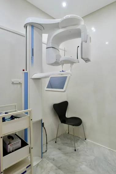 A modern dental X-ray and imaging room featuring a white and blue panoramic dental imaging machine, a digital touch screen, a black chair, and a medical supply cart on marble flooring.