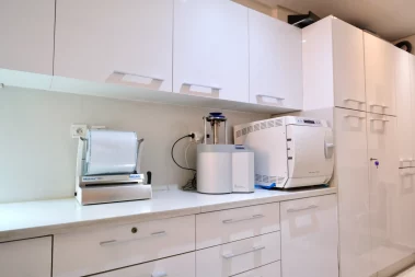 A modern medical or dental sterilization room featuring white cabinetry, a counter top with a sealing machine, an automated hand piece care unit, and an autoclave machine.