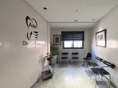A modern dental clinic waiting room featuring white walls with the "AM Dental Care" logo, black and chrome seating, a window with dark blinds, and marble flooring.