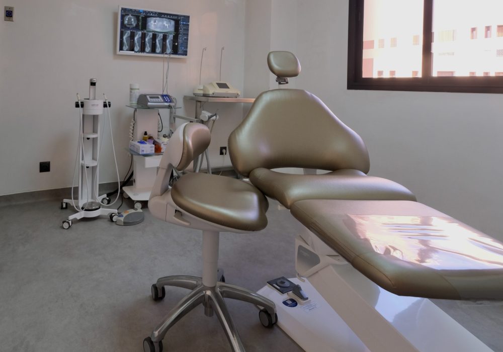 Dental treatment room with a champagne-colored reclining chair, adjustable stool, and a wall-mounted screen displaying dental X-ray scans.